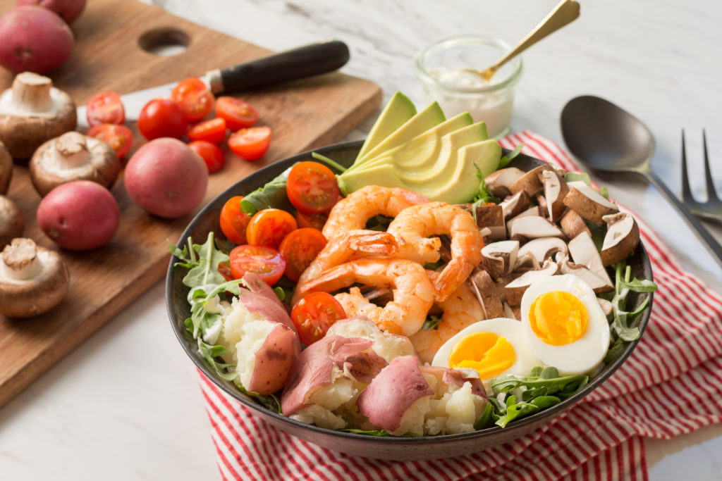 Bowl of shrimp cobb salad next to cutting board of vegetables.