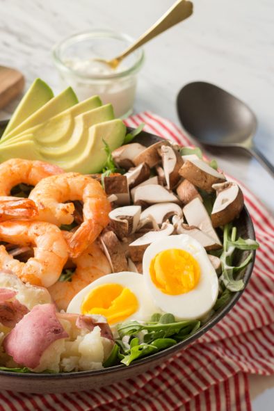 Bowl of shrimp cobb salad next to cutting board of vegetables.