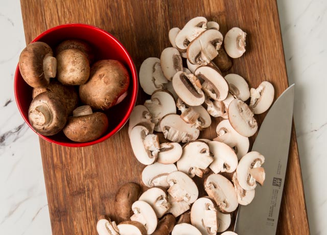 Sliced crimini mushrooms on a cutting board next to small bowl of whole mushrooms.