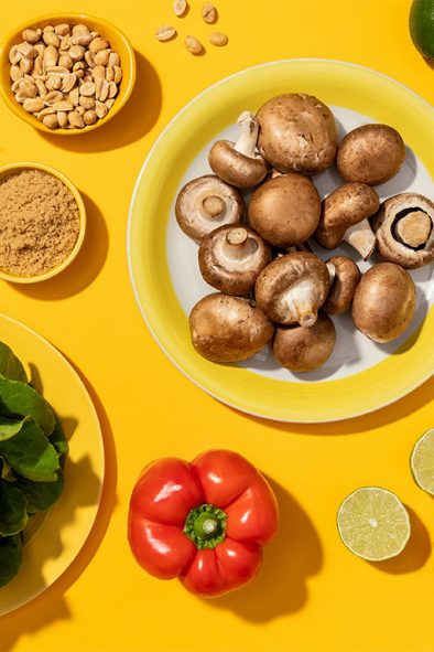An assortment of fresh cooking ingredients arranged on a yellow surface, featuring mushrooms, leafy greens, carrots, bell pepper, limes, and green onions.