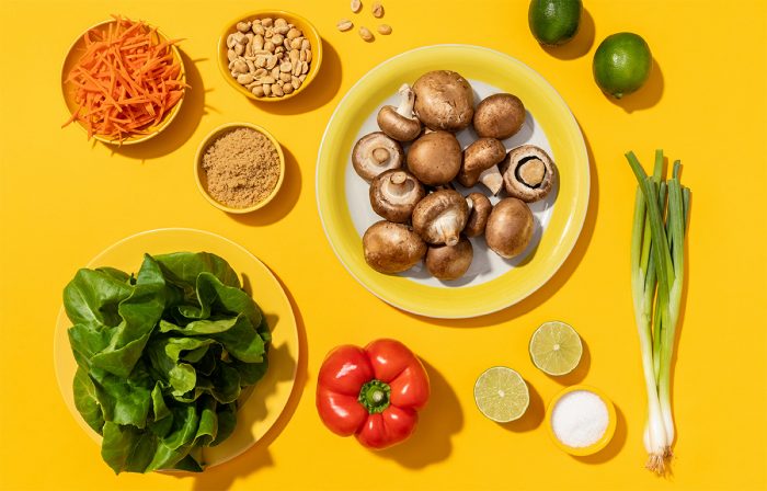An assortment of fresh cooking ingredients arranged on a yellow surface, featuring mushrooms, leafy greens, carrots, bell pepper, limes, and green onions.