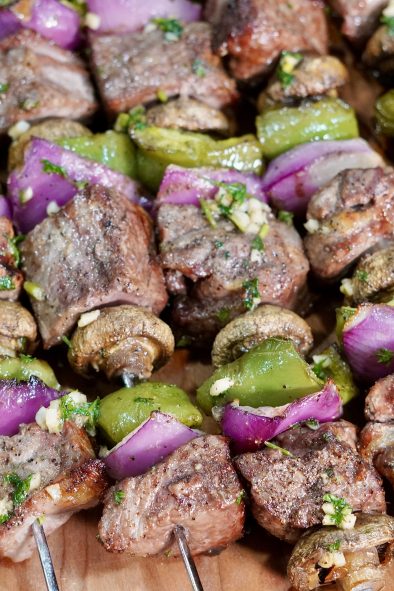 A close-up shot of several grilled beef and vegetable skewers arranged on a wooden cutting board. Each skewer features alternating pieces of browned steak, whole white button mushrooms, vibrant green bell pepper chunks, and bright purple red onion pieces. Some pieces of steak and vegetables show a light green garnish of what appears to be chopped parsley. The focus is sharp on the foreground skewers, with the background slightly blurred.