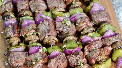 A close-up shot of several grilled beef and vegetable skewers arranged on a wooden cutting board. Each skewer features alternating pieces of browned steak, whole white button mushrooms, vibrant green bell pepper chunks, and bright purple red onion pieces. Some pieces of steak and vegetables show a light green garnish of what appears to be chopped parsley. The focus is sharp on the foreground skewers, with the background slightly blurred.