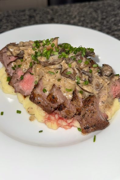 A medium-rare steak, sliced and fanned out over a bed of mashed potatoes, is generously covered with a creamy mushroom sauce and sprinkled with chopped chives, presented on a white plate.