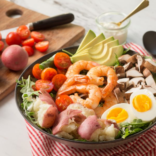 A healthy bowl contains shrimp, avocado, hard-boiled eggs, cherry tomatoes, mushrooms, and red-skinned potatoes on a bed of greens.