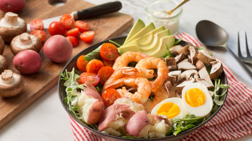 A healthy bowl contains shrimp, avocado, hard-boiled eggs, cherry tomatoes, mushrooms, and red-skinned potatoes on a bed of greens.