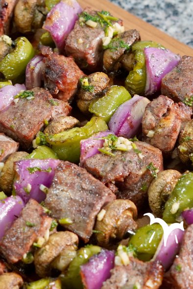 A close-up shot of several grilled beef and vegetable skewers arranged on a wooden cutting board. Each skewer features alternating pieces of browned steak, whole white button mushrooms, vibrant green bell pepper chunks, and bright purple red onion pieces. Some pieces of steak and vegetables show a light green garnish of what appears to be chopped parsley. The focus is sharp on the foreground skewers, with the background slightly blurred.