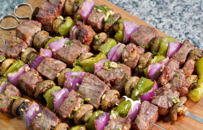 A close-up shot of several grilled beef and vegetable skewers arranged on a wooden cutting board. Each skewer features alternating pieces of browned steak, whole white button mushrooms, vibrant green bell pepper chunks, and bright purple red onion pieces. Some pieces of steak and vegetables show a light green garnish of what appears to be chopped parsley. The focus is sharp on the foreground skewers, with the background slightly blurred.