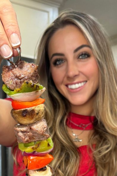 Katherine Salom smiles while holding up a grilled skewer filled with steak, mushrooms, and colorful vegetables.