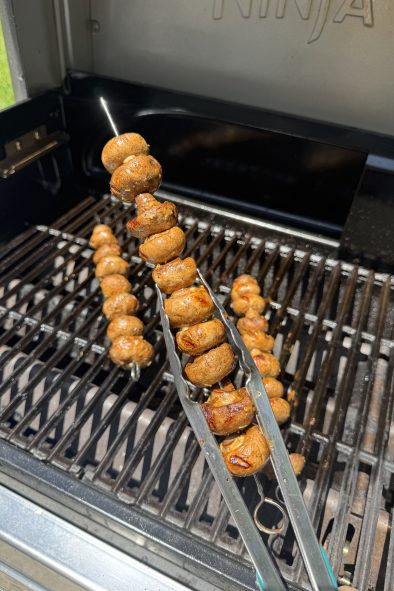 Tongs hold a skewer of grilled white button mushrooms over an outdoor gas grill with two other skewers on the grates.
