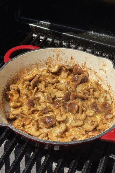 A red skillet of sautéed shiitake mushrooms sits on a grill grate.