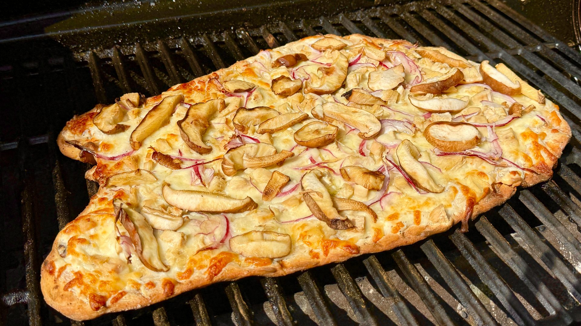 A rectangular flatbread pizza, topped with melted cheese, thinly sliced red onion, and shiitake mushrooms, is cooking on the grates of a black outdoor grill. The crust is golden brown and slightly charred in spots.